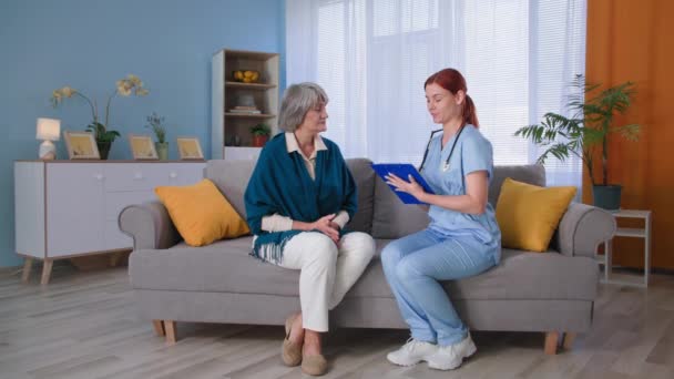 Ιατρική επίσκεψη στο σπίτι, νεαρή γυναίκα νοσοκόμα με στολή με πρόχειρο συμβουλεύει ηλικιωμένη γυναίκα ασθενή στον καναπέ — Αρχείο Βίντεο
