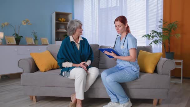 Badania lekarskie w domu, lekarka w mundurze ze schowkiem konsultingowym starsza pani pacjentka na kanapie — Wideo stockowe