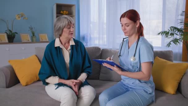 Portrét lékaře a starší pacientky, terapeutka v uniformě si dělá poznámky do schránky, zatímco doma konzultuje starší ženu — Stock video