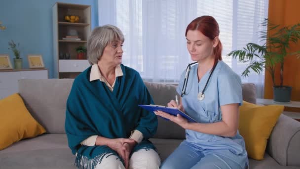 Hälso- och sjukvård, ung läkare i uniform lyssnar på symptom på äldre patient och antecknar på Urklipp sitter på soffan — Stockvideo