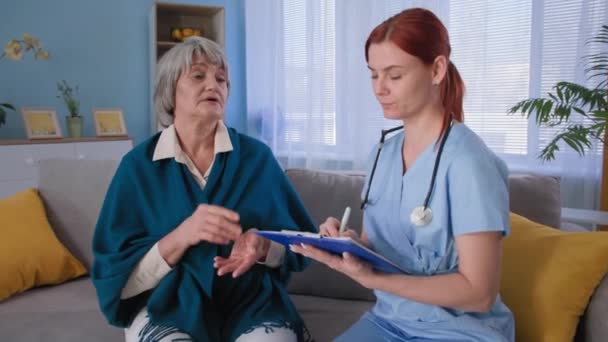 Dokter muda berkonsultasi dengan wanita dewasa yang sakit dan mencatat di papan klip, terapis perempuan berseragam mendengarkan gejala-gejala pasien senior selama kunjungan ke rumah — Stok Video