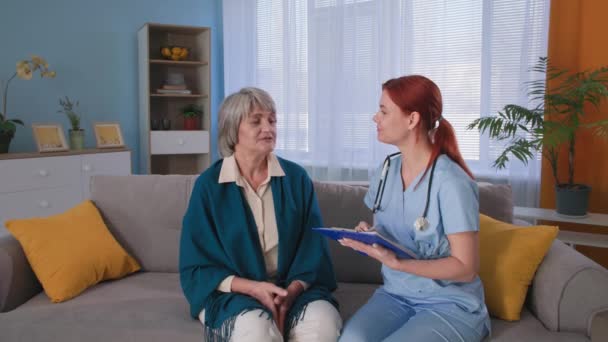 Terapeut v uniformě konzultuje seniorskou pacientku při domácí návštěvě, portrét šedovlasé babičky s lékařkou dívající se do kamery — Stock video
