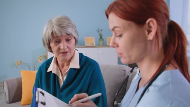 Nagymama beszél a betegségeiről az orvosnak otthon, női terapeuta jegyzetel az írótáblán, és figyelmesen hallgatja a beteget — Stock videók