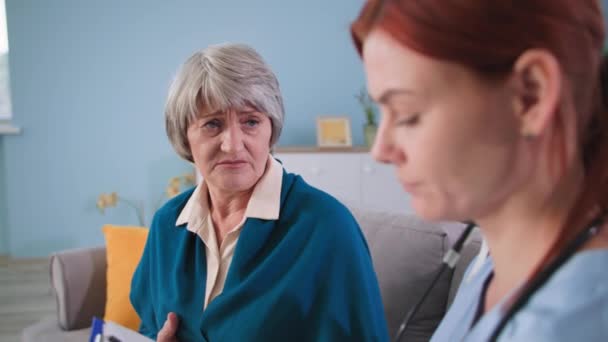 Jovem enfermeira se comunica com paciente do sexo feminino na sala de estar, terapeuta faz anotações na área de transferência e escuta a idosa em casa — Vídeo de Stock
