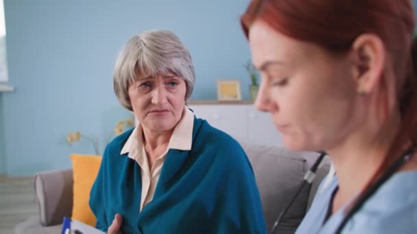 Junge Krankenschwester kommuniziert mit Patientin im Wohnzimmer, Therapeutin macht Notizen auf Klemmbrett und hört der Seniorin zu Hause zu — Stockvideo