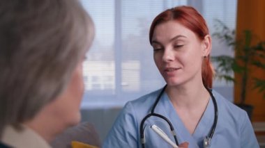 Yaşlı bir hastayla konuşan profesyonel bir kadın terapist. Steteskopla mavi üniformalı aile doktoru. Muayene sırasında notlar alıyor.
