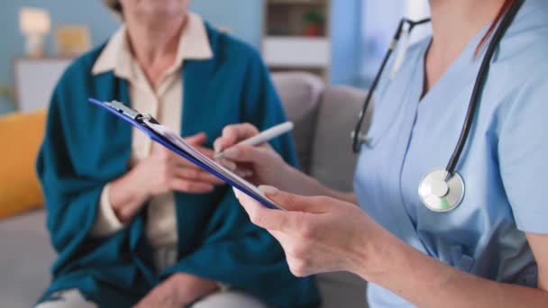 Handen van arts met pen maakt notities op klembord tijdens een consult met vrouwelijke patiënt, therapeut in uniform met een stethoscoop praat over de behandeling thuis, close-up — Stockvideo
