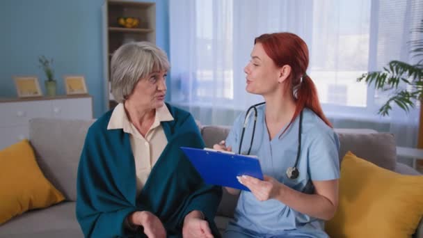 Uśmiechnięta pielęgniarka przytula ramiona szczęśliwej pacjentki i patrzy w kamerę, miły lekarz robi notatki na tablicy i rozmawia ze starszą kobietą w domu — Wideo stockowe