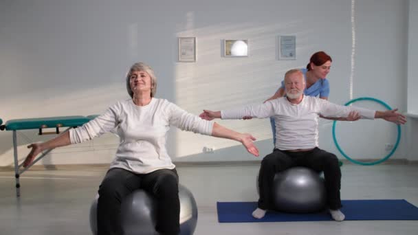 Пара пожилых пациентов, выполняющих упражнения, сидя на фитболах, медсестра, помогающая бабушке и дедушке поднять руки во время тренировки в офисе — стоковое видео