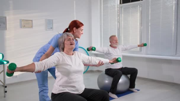 Glada äldre par som sitter på träningsbollar räcka upp handen med hantlar medan du tränar tillsammans med sjuksköterskan hemma — Stockvideo
