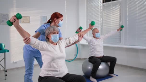 Para w podeszłym wieku ćwicząca na piłeczkach gimnastycznych ze specjalistą od mięśni w szpitalu podczas pandemii, staruszka podnosi ramiona przy wsparciu pielęgniarki, zamaskowane osoby — Wideo stockowe