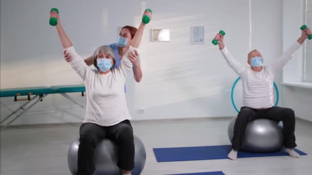 Zdrowie i kontrola ciała, starsza para w maskach medycznych wykonująca ćwiczenia z hantlami siedzącymi na piłeczkach fitballowych, pielęgniarka pomagająca emerytowi podnieść ręce podczas treningu — Wideo stockowe