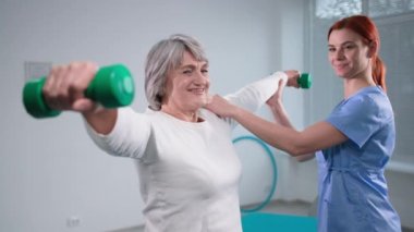 Rehabilitasyon ve tedavi, genç fizyoterapist yaşlı kadınların ellerini uzatıyor, halterli, iyileşme sürecinde.