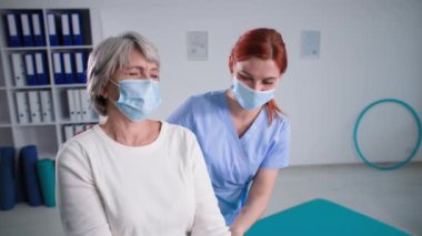 Covid 19 sırasında evde sağlık hizmeti, tıbbi maskeli yaşlı kadın, fizyoterapist halterli aktif büyükanneye yardım ediyor.