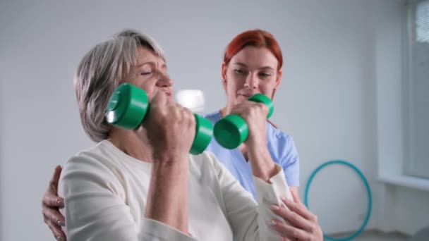 Ženský lékař v domově důchodců pomáhá seniorce zotavit, šťastný šedovlasý důchodce protahuje paže s činkami — Stock video