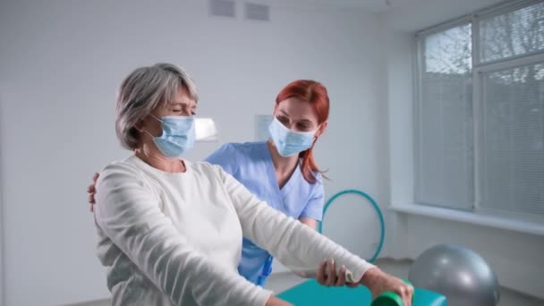 Pensioner goes in for sports and healthy lifestyle in nursing home, old woman lifts dumbbells with help physiotherapist sitting on couch — Stock Video