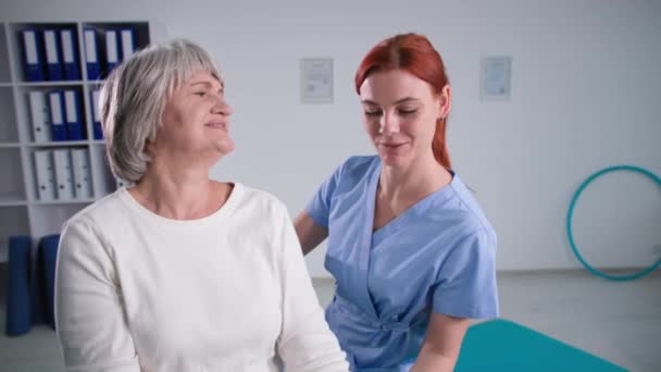 Alte Frau trainiert zu Hause mit Krankenschwester in Uniform, glückliche Großmutter beugt Arme mit Hanteln und blickt in die Kamera in der Reha — Stockvideo