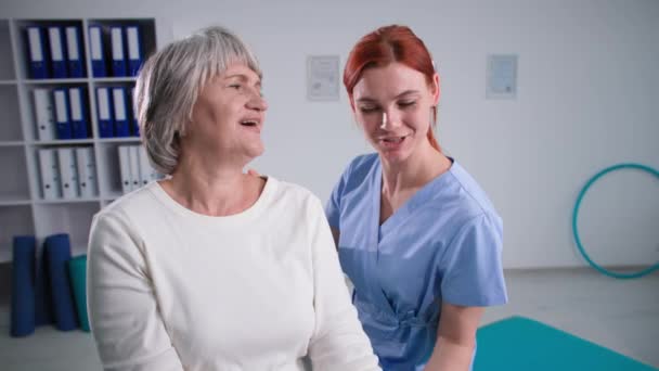 Фитнес-упражнения для пожилых людей в номере, услуги медсестры, помогающей активной пожилой женщине накачивать мышцы плеча гантелями — стоковое видео