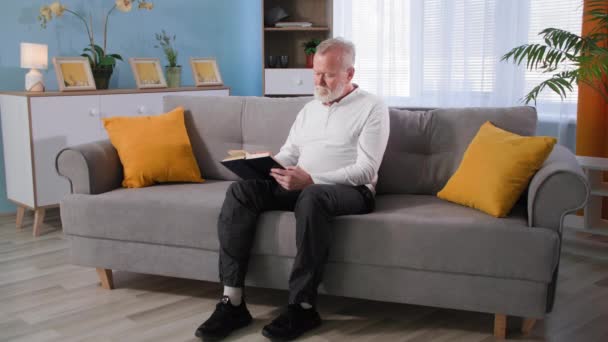 Senior old gray-haired man reads a book and then slowly stand up from the sofa in living room, grandfather suffering from knee pain at home — Stock Video