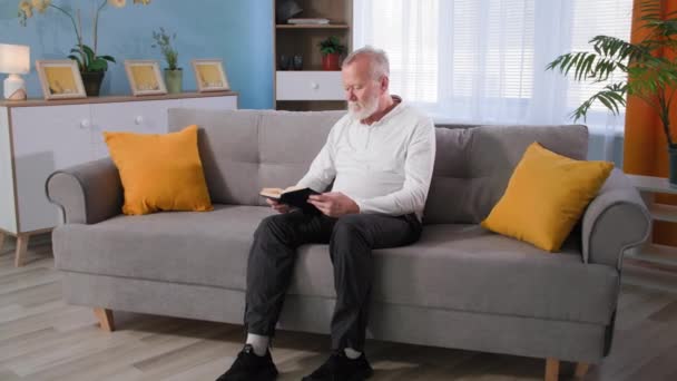 Old man reads book then gets up from the couch and feels pain in his leg, granddad with osteoporosis rubs his knee at home — Stock Video