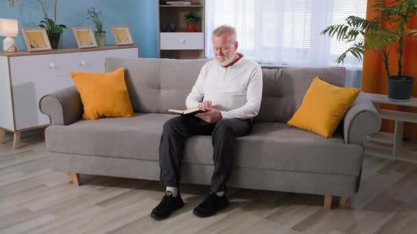 Tempo libero in pensione, anziano uomo dai capelli grigi gira pagine durante la lettura del libro mentre seduto sul divano a casa — Video Stock