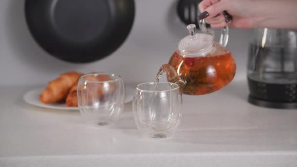 Mujer joven vierte té caliente de hierbas de la tetera de vidrio en tazas transparentes durante el desayuno de la mañana en la cocina — Vídeos de Stock