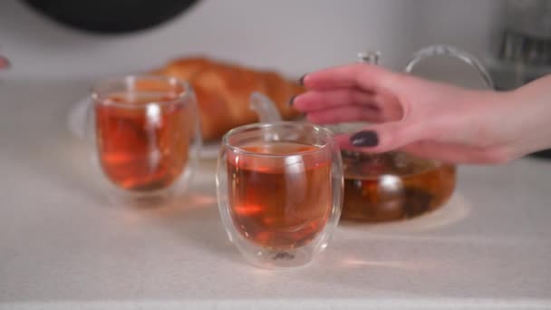 Naturaleza muerta, manos femeninas toman tazas transparentes con delicioso té de hierbas saludables contra el fondo de una tetera con una bebida caliente y croissants, desayuno — Vídeo de stock