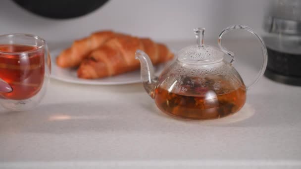 Estilo de vida comida, as mãos femininas colocar copos transparentes com chá de ervas quente na mesa contra o fundo de um bule de chá e croissants na cozinha — Vídeo de Stock