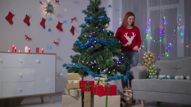Fiatal nő készül az újévi ünnepekre, lány piros pulóverben díszíti a karácsonyfát lógó golyók mesterséges ágak, egy csomó ajándék dobozok a fa alatt — Stock videók