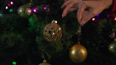 Yeni yıl atmosferi, kadın elinin yakın çekimi küçük çanlar, toplar ve çelenklerle bir Noel ağacını süslüyor.