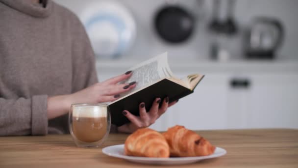 Ruhe zu Hause, junge Frau liest Buch und trinkt leckeren Kaffee mit Milch und Croissants in der Küche am Tisch — Stockvideo