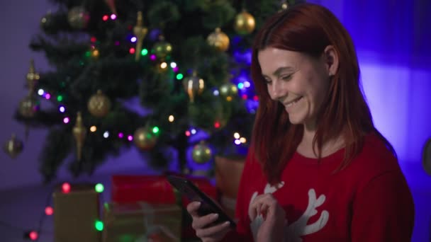 Happy girl with mobile phone on new years eve, young woman shows at the camera a smartphone with a green screen and a gift on the background of a Christmas tree with many lights at night — Stok video