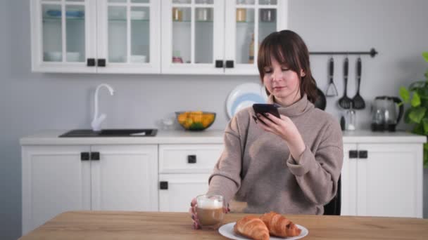 Hvile hjemme tar unge kvinner bilde av frokost på mobiltelefon for virtuelle venner og drikker kaffe med melk og croissanter mens de sitter ved bordet på kjøkkenet – stockvideo