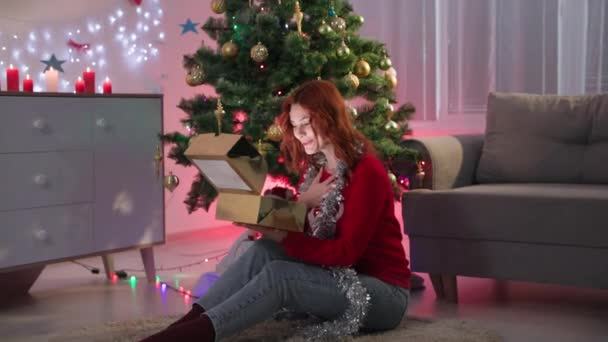 New year gift, happy young beautiful woman in a red sweater opens box at night time, light shines on her face on the background of christmas tree with garland — Stockvideo