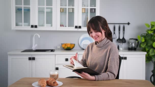 Спосіб життя, молода жінка з книгою в руках читає і п'є каву, сидячи вдома за столом на фоні кухні — стокове відео