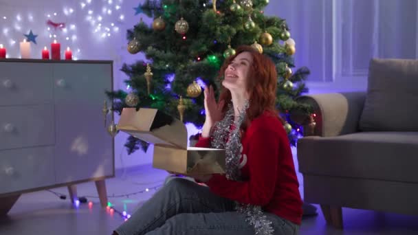 Happy new year, girl in red sweater opens gift box with magic light surprised and laughs at dark on background christmas tree — Stok video