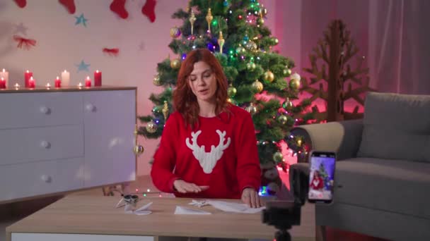 Blog about xmas decor, girl blogger teaches how to make a snowflake out of paper in front of camera of smartphone sitting at the table at home on the background of a Christmas tree — Stok video