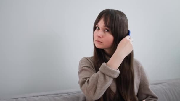 Красивая молодая женщина расчесывает ухоженные длинные волосы расческой, сидя дома на диване — стоковое видео