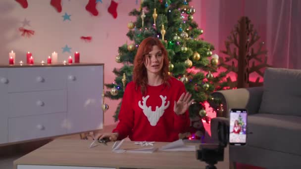 Leisure on new year eve, blogger girl demonstrates on a mobile phone camera how to cut snowflake out of paper using scissors sitting on the background of a Christmas tree — Stok video