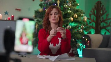 young woman takes off new year blog near Christmas tree, girl with scissors cuts out the decor from paper and then opens a snowflake in front of mobile phone camera