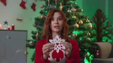 online training on how to make paper snowflakes, young woman blogger talks about crafts for New Year interior while sitting at home near a Christmas tree, webcam view