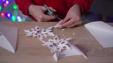 christmas decor, close-up of female hands with scissors cuts snowflake out of paper while sitting at the table at home