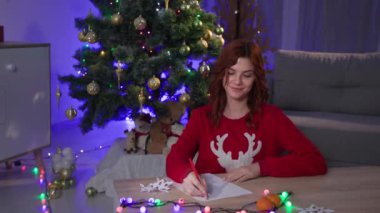 letter to Santa Claus, happy dreamy girl writes a wish list for the new year and then lays down note near the christmas tree in a cozy dark room with a festive interior