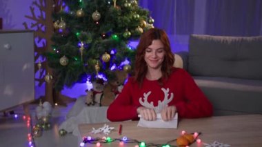 spirit of christmas, girl in red sweater puts a letter with wishes for gift near Xmas tree to Santa Claus, portrait of happy dreamy young woman in cozy dark room
