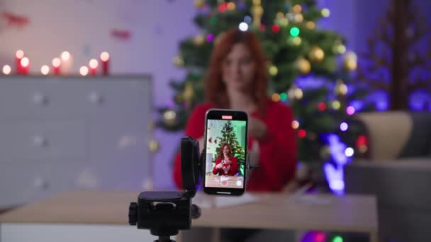 Mobile phone on tripod takes off new year blog, girl with scissors shows how to make snowflake out of paper while communicating with subscribers on background of Christmas tree — 비디오