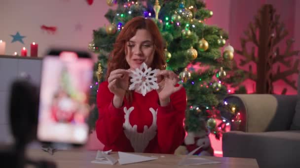 Christmas crafts, female blogger remotely on camera of mobile phone tells how to make paper snowflake for new year near xmas tree — Vídeo de Stock