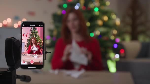 Mobile phone on tripod in focus, female blogger shows how to make snowflake out of paper while communicating with subscribers on camera near Christmas tree — Vídeo de Stock