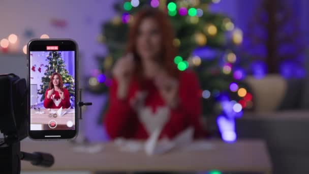 Smartphone on tripod in focus, female blogger shows how to make paper craft then throws snowflakes in air on camera near xmas tree — Vídeo de Stock
