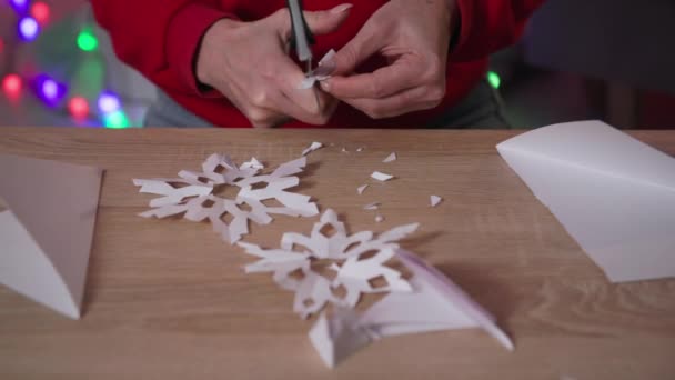 New Year decor, close-up of female arms with scissors cuts snowflake out of paper and then opens it while sitting at the table at home — стоковое видео