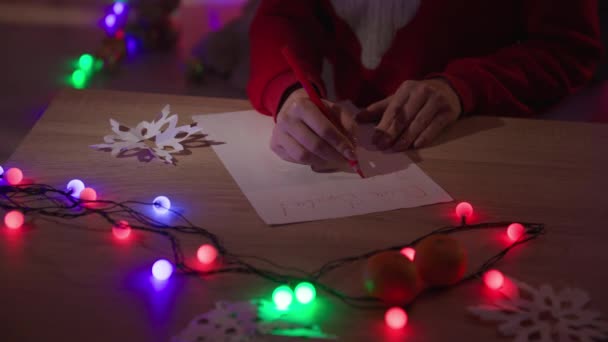 Merry Christmas and New Year, woman with a red pencil write letter with wishes for Santa Claus while sitting at a table with garland in a cozy dark room — стоковое видео